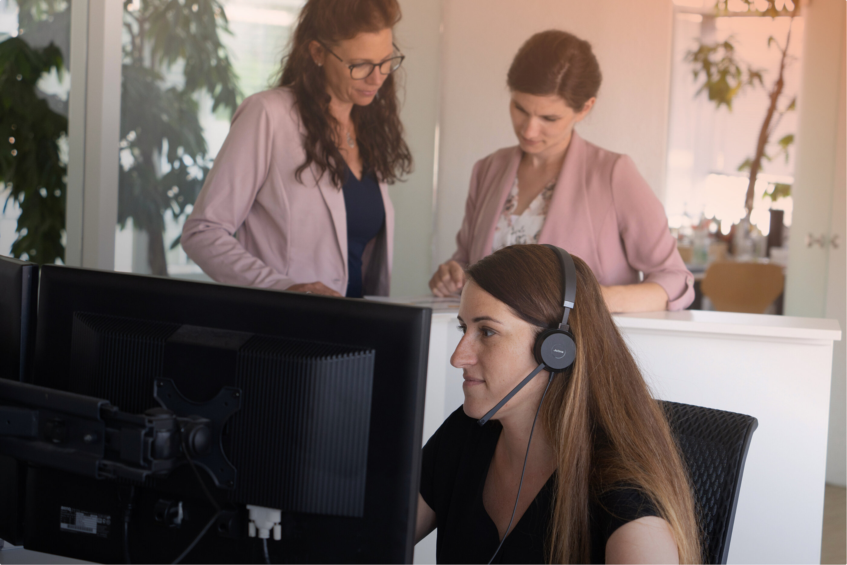 Im Vordergrund sitzt eine Frau mit Headset arbeitend am PC, während zwei Frauen im Hintergrund etwas besprechen