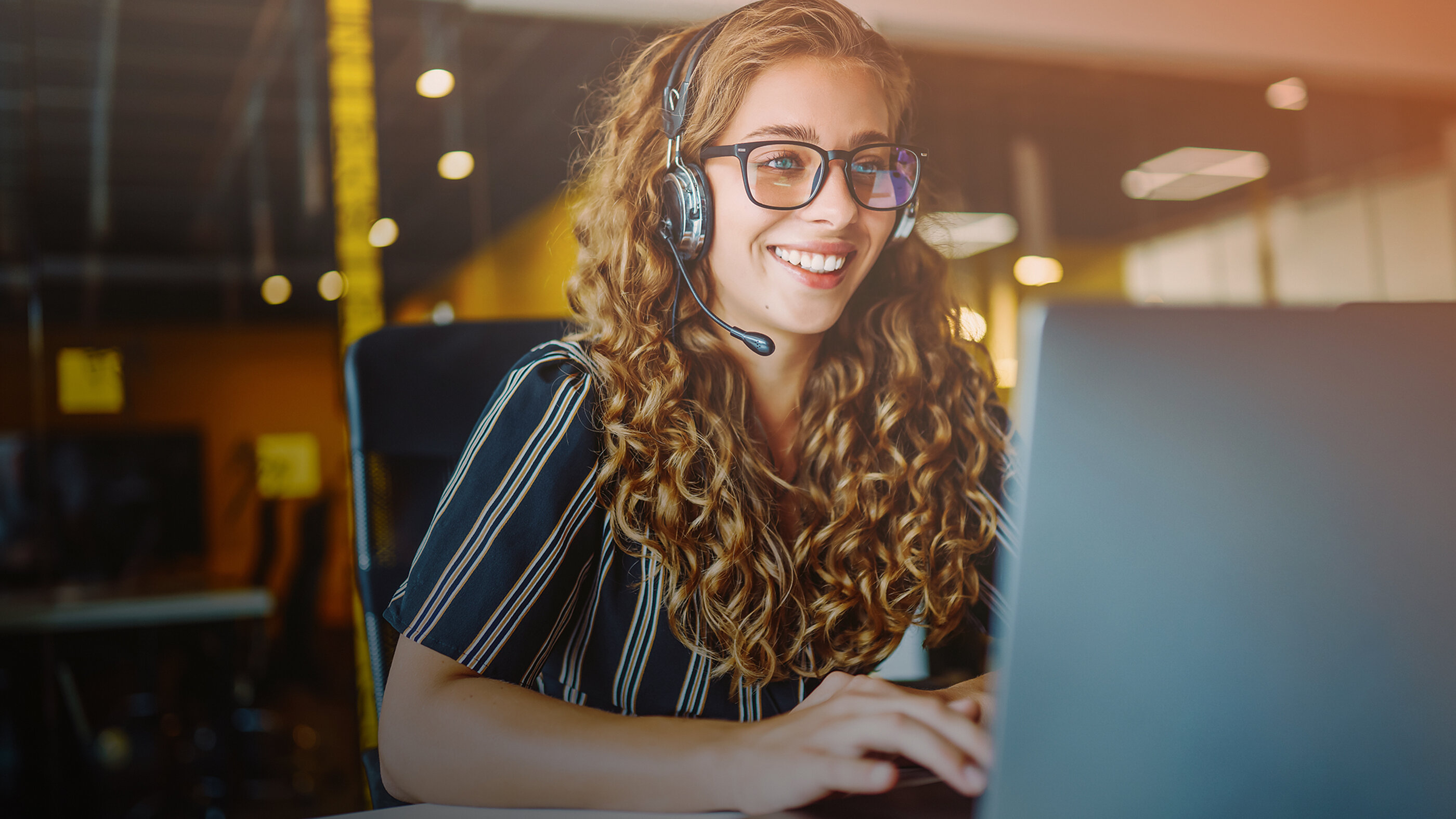 Lächelnde Mitarbeiterin mit Headset und Brille am Computer im Kundenservice